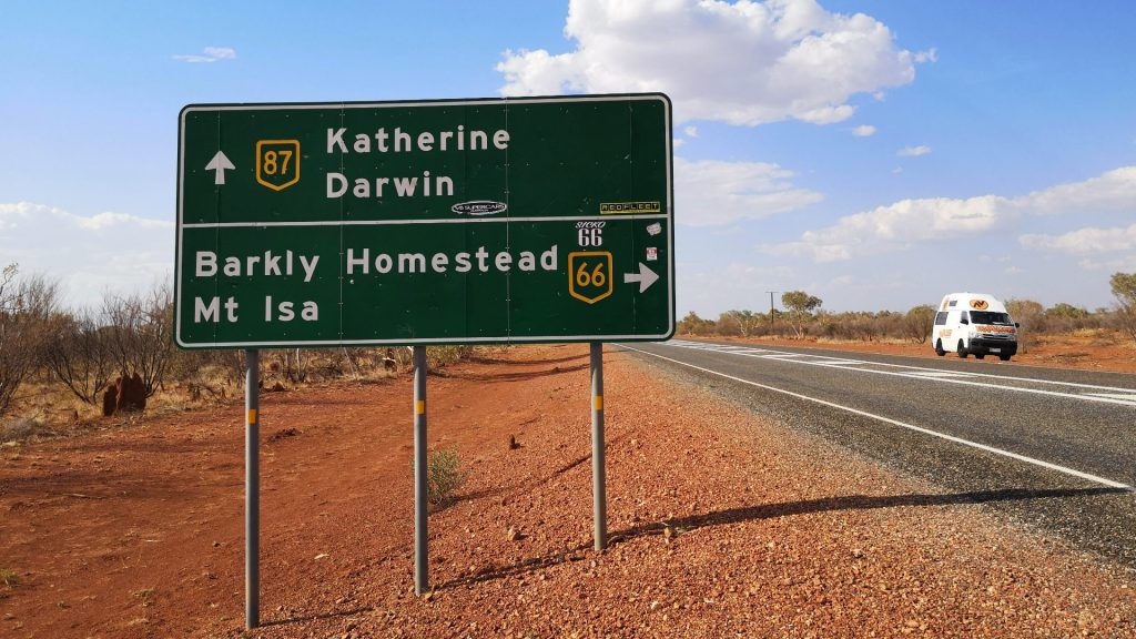 Road sign pointing to Katherine, Darwin and Mount Isa on red soil beside a black road.