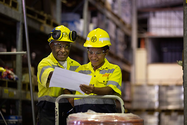 Two female Ok Tedi engineers in yellow high vis, standing in a warehouse, reviewing something on a piece of paper.
