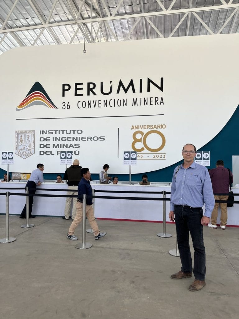 Man in black trousers and blue button up shirt standing in front of the registration desk. Behind the desk is a large sign saying 'PERUMIN'.