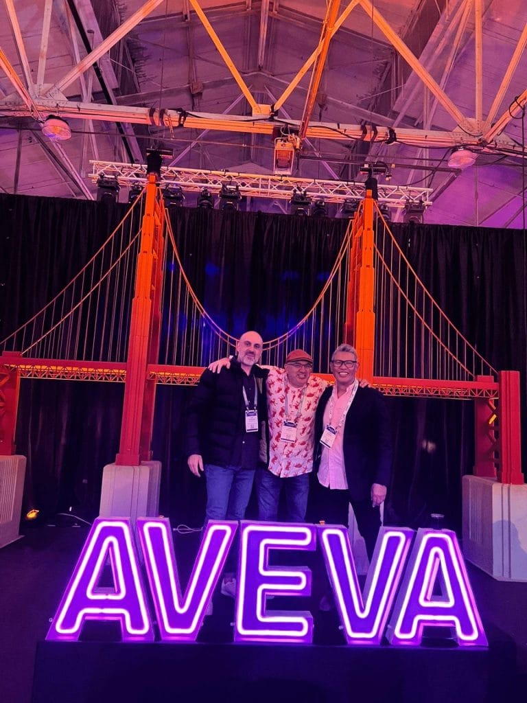 Three men standing behind a lit up, purple AVEVA sign, with a background of a fake Golden Gate Bridge.