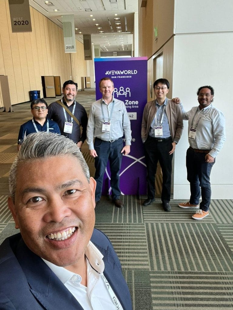 Man taking a selfie with a group of other men and a purple AVEVA banner in the background.