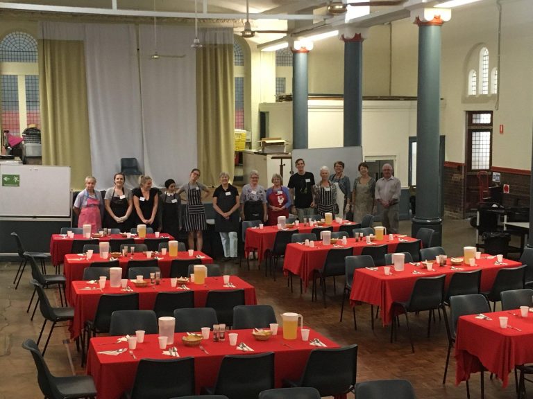 A number of volunteers stand in a row at the back of a large room set with tables covered in red table clothes and crockery/cutlery, ready to serve people.