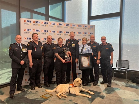 8 volunteers and a labrador stand/lie in front of a background, holding awards.