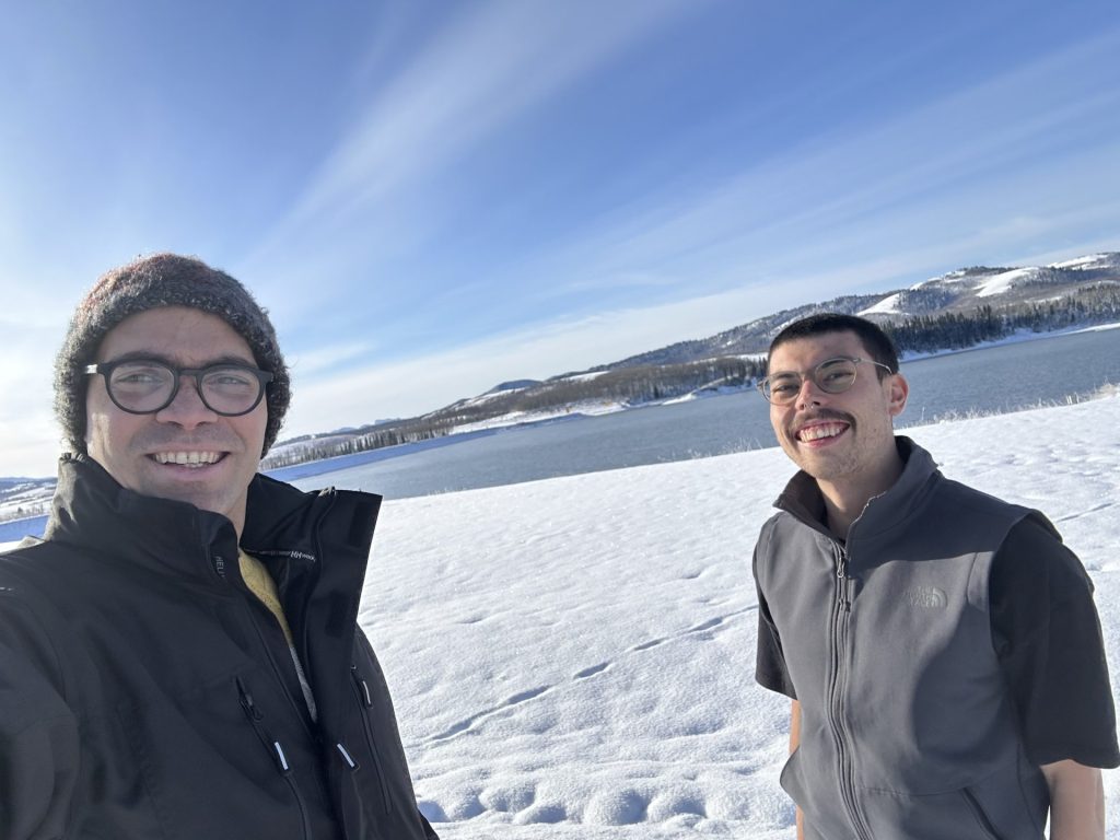 Two men smiling into the camera with a snowy background and bright blue sky
