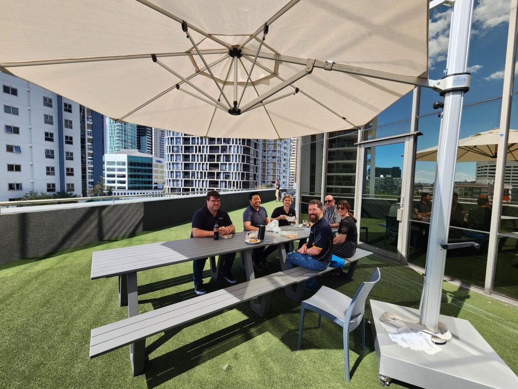 Graduate engineer Jake Minehan and colleagues sitting together at an outdoor table, on the terrace of a high-rise building, eating lunch under the shade of an umbrella