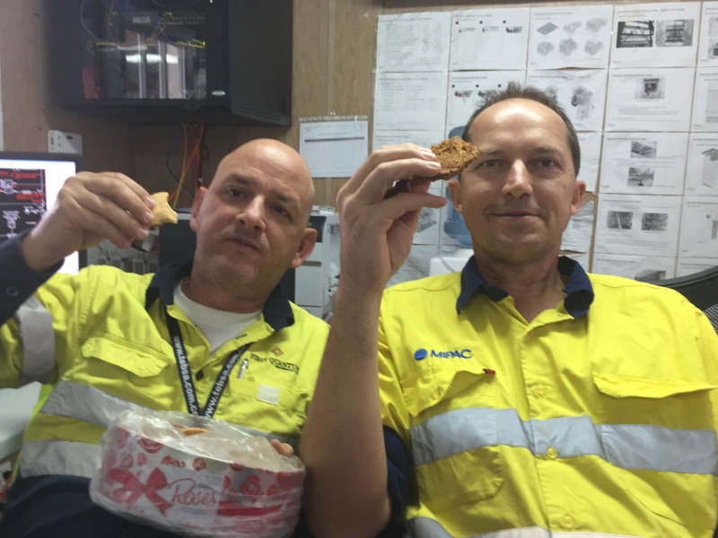 Two men in high visibility shirts sitting in a mine site office holding up a cookie each from a tin.