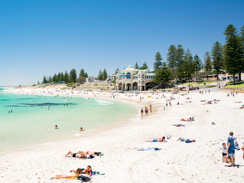 Picture of Cottesloe Beach in Perth, Western Australia on a sunny day. Picture shows white sand, people lying in sand and a historical building in the background surrounded by pine trees.