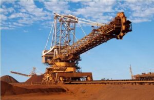 a photo of an iron ore reclaimer against a blue, cloud-spotted sky with red earth below.