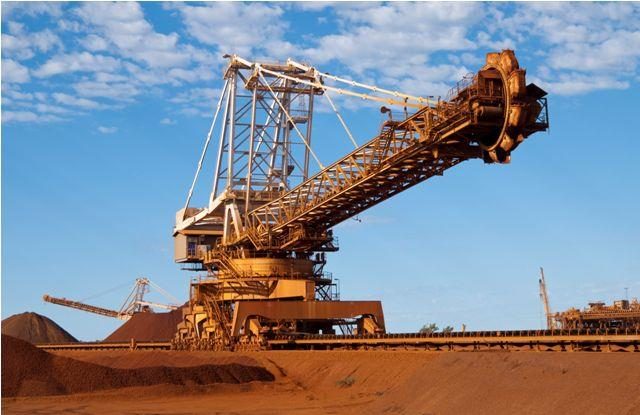 a photo of an iron ore reclaimer against a blue, cloud-spotted sky with red earth below.