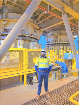 An engineer in yellow and blue high vis posing for a photo in front of some mining machinery that is also yellow and grey.
