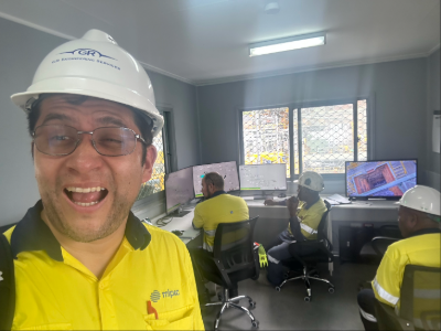 A man in a yellow hi-vis shirt and white safety helmet (a Mipac engineer) grins at the camera. In the background are three people sitting in a small office, working on computers. Through the windows, you can just make out a mine site.